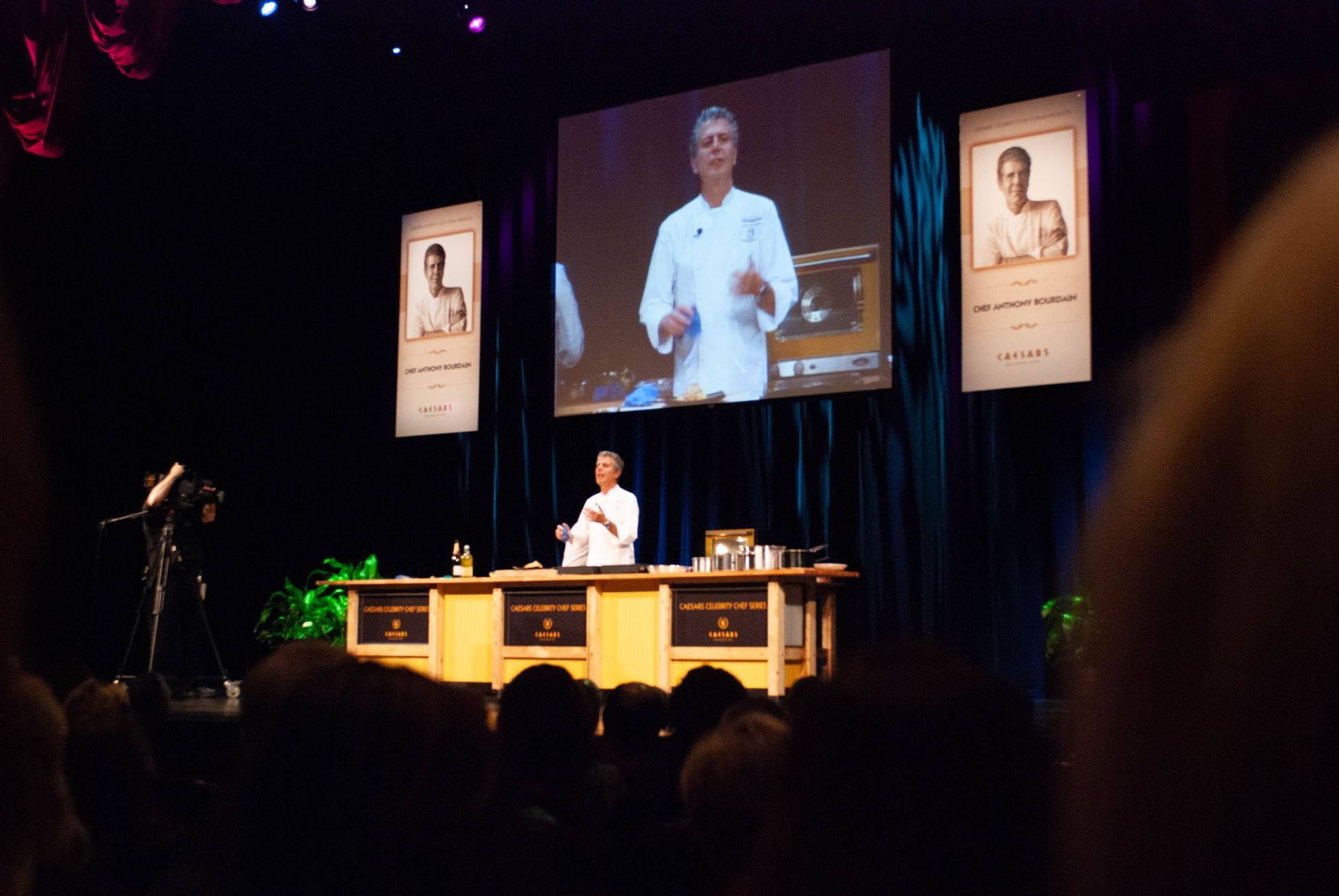 Speaker on stage in a dark auditorium with projected screen and banners, addressing an audience from a desk