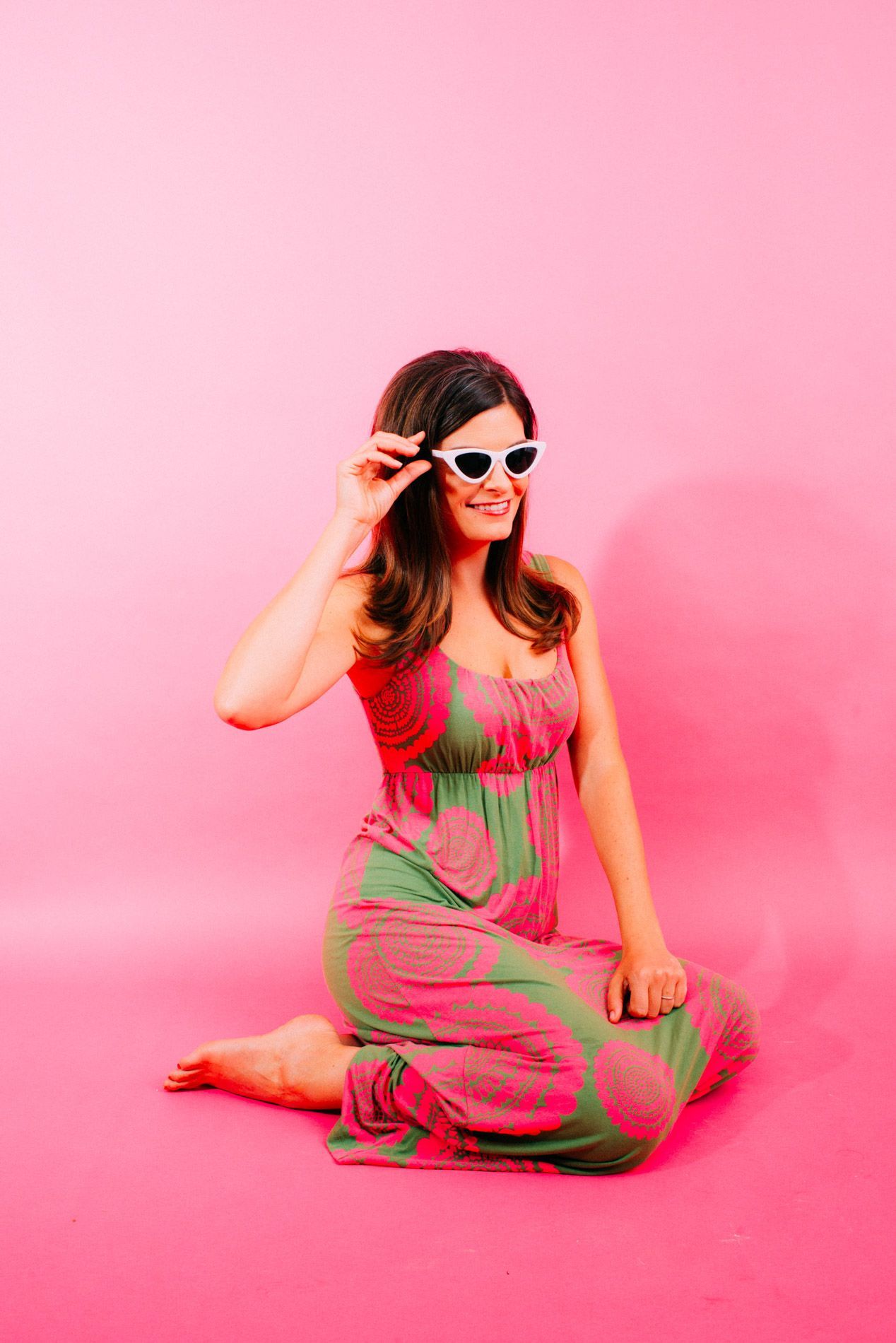 Person kneeling on a pink floor against a pink backdrop, adjusting sunglasses in a green-and-pink outfit