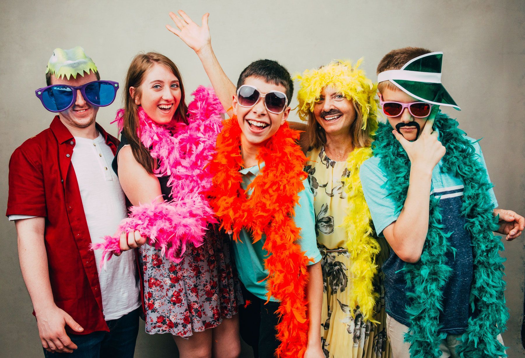 Five people in colorful feather boas and sunglasses posing together against a plain wall.