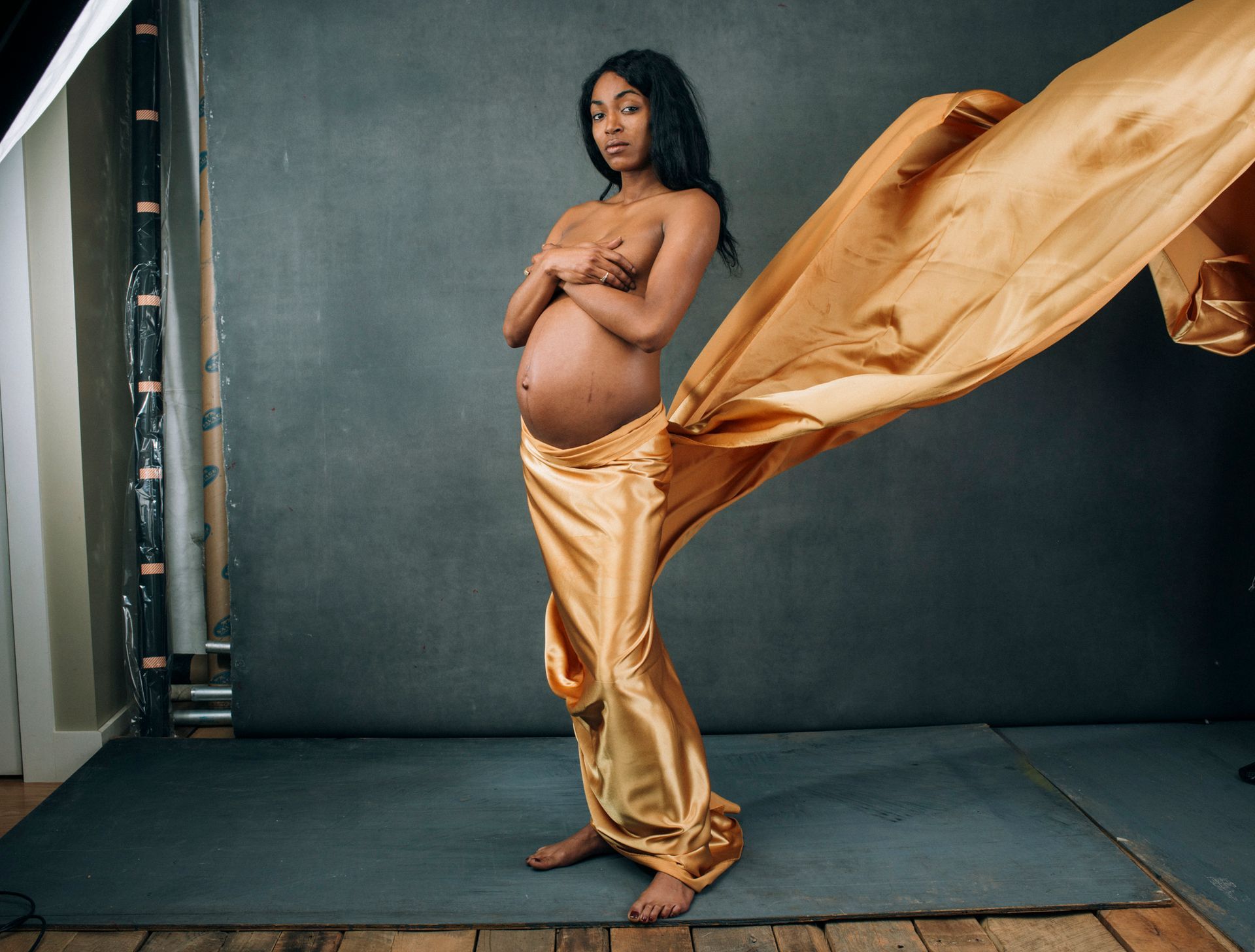 Pregnant mother in a gold draped fabric pose against a dark studio backdrop, fabric billowing beside them, photographed by asheville maternity photographer sandlin gaither 
