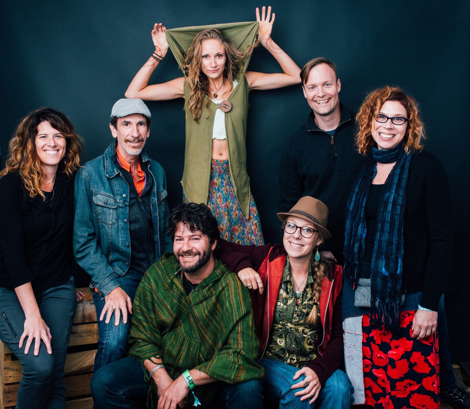 Seven people pose smiling against a dark backdrop, with one person standing behind the others with raised hands.