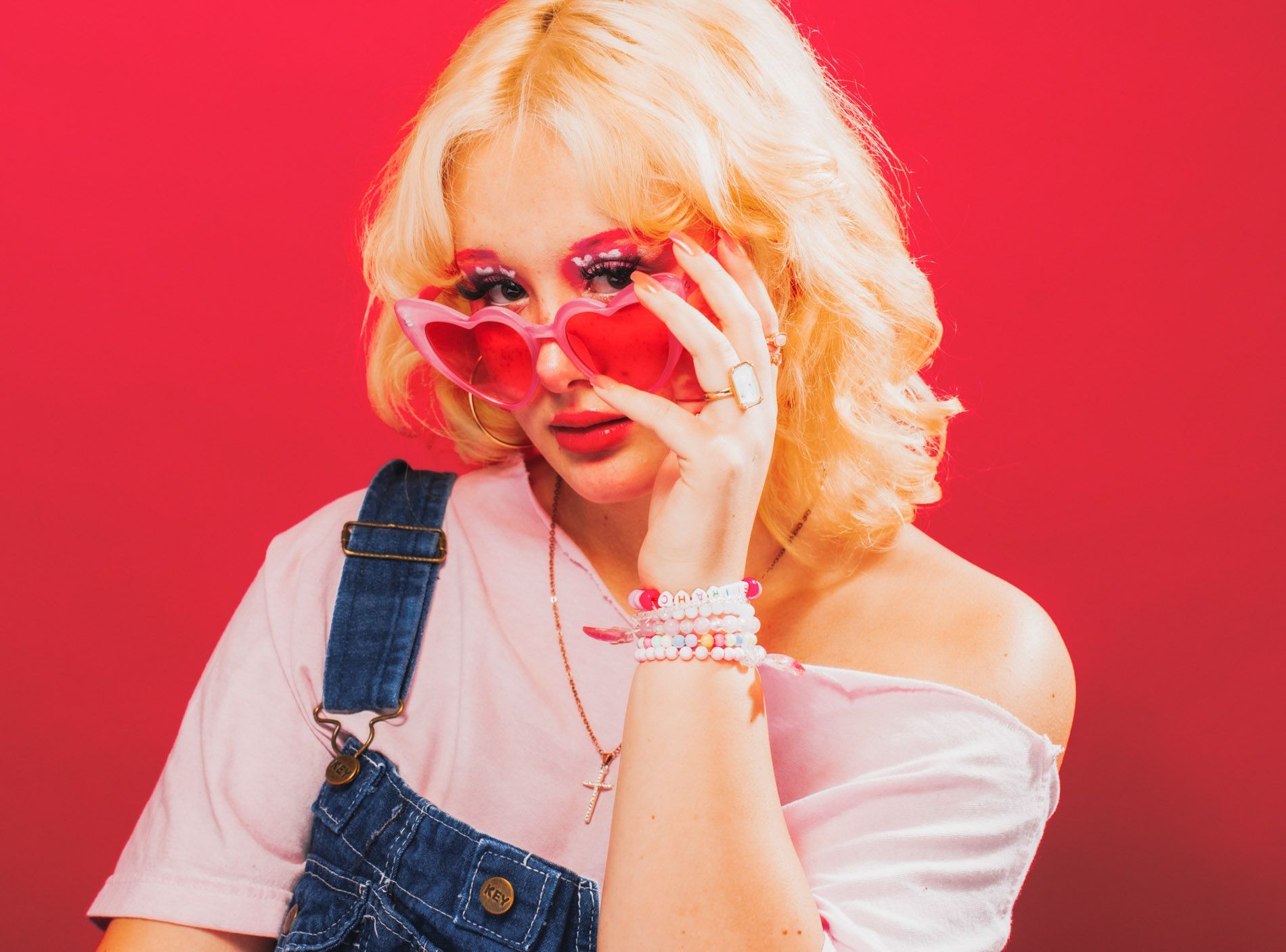 Blonde person in pink sunglasses and denim overalls posing against a bright pink background, portrait in studio by Asheville photographer Sandlin Gaither
