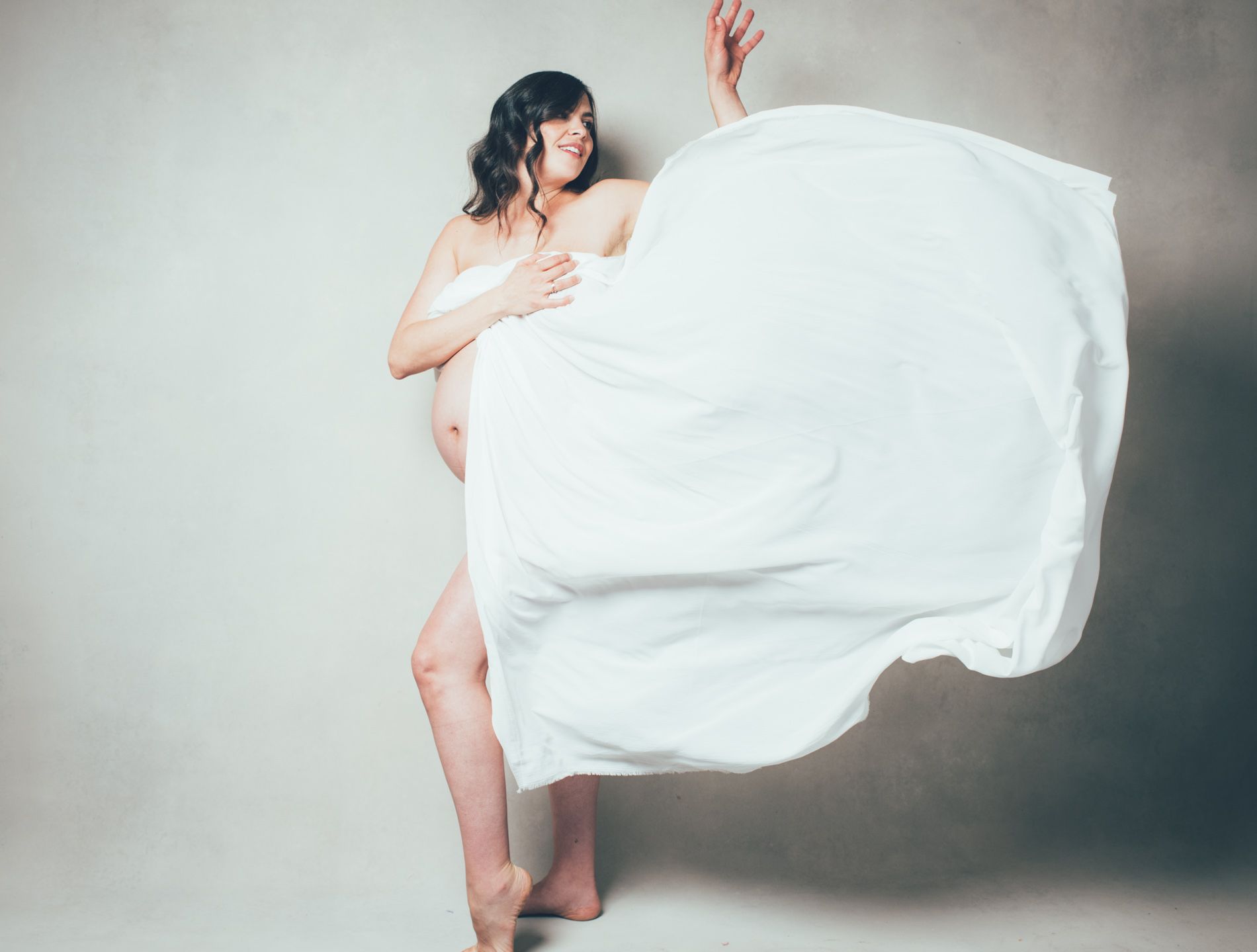 Pregnant mother in white fabric posing with flowing drape against a plain gray background, photographed in ahseville by maternity photographer Sandlin Gaither