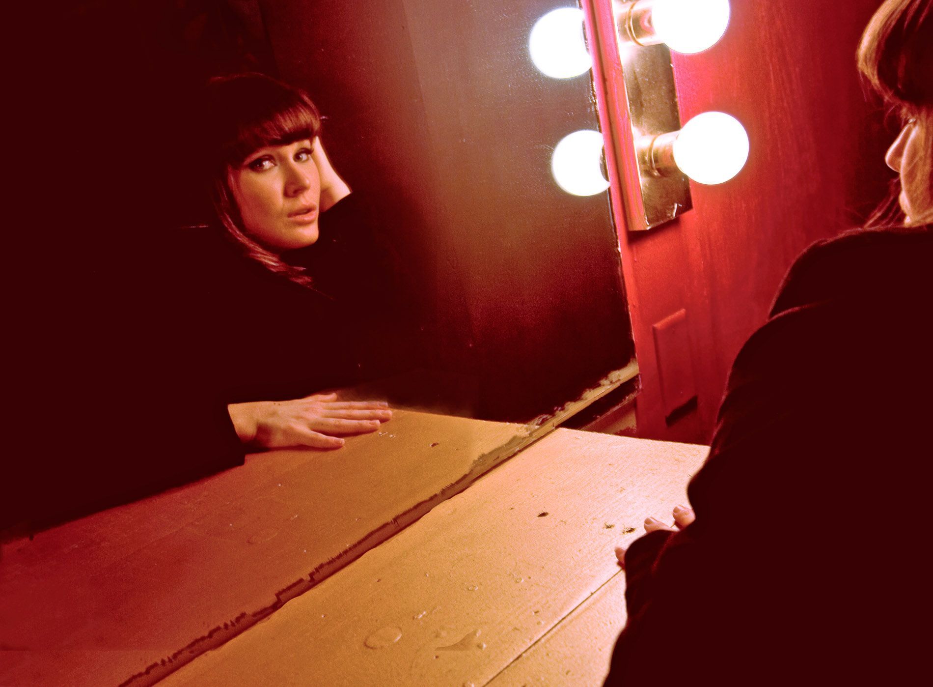 Person seated at a vanity mirror with glowing bulbs in a red-lit dressing room