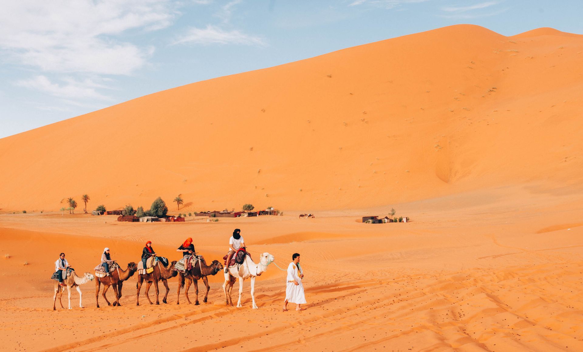 Camels with riders crossing orange desert dunes under a bright sky