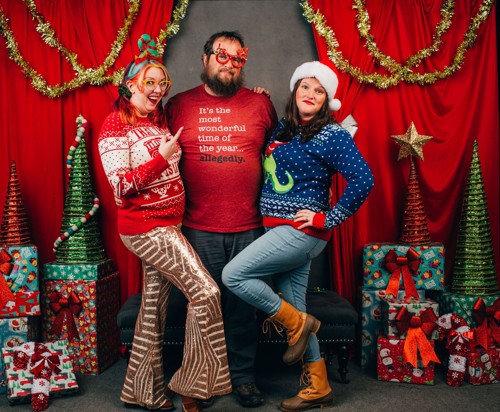 Three people posing in festive holiday sweaters before a red Christmas backdrop with gifts and garlands