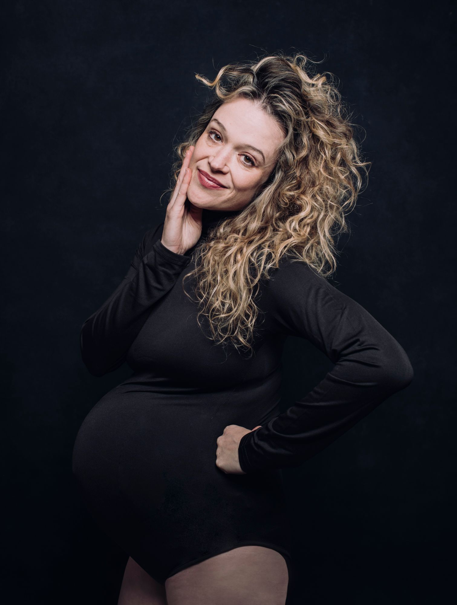 Pregnant woman in a black dress posing against a dark background, smiling with one hand on her cheek, photographed by asheville maternity photographer sandlin gaither 
