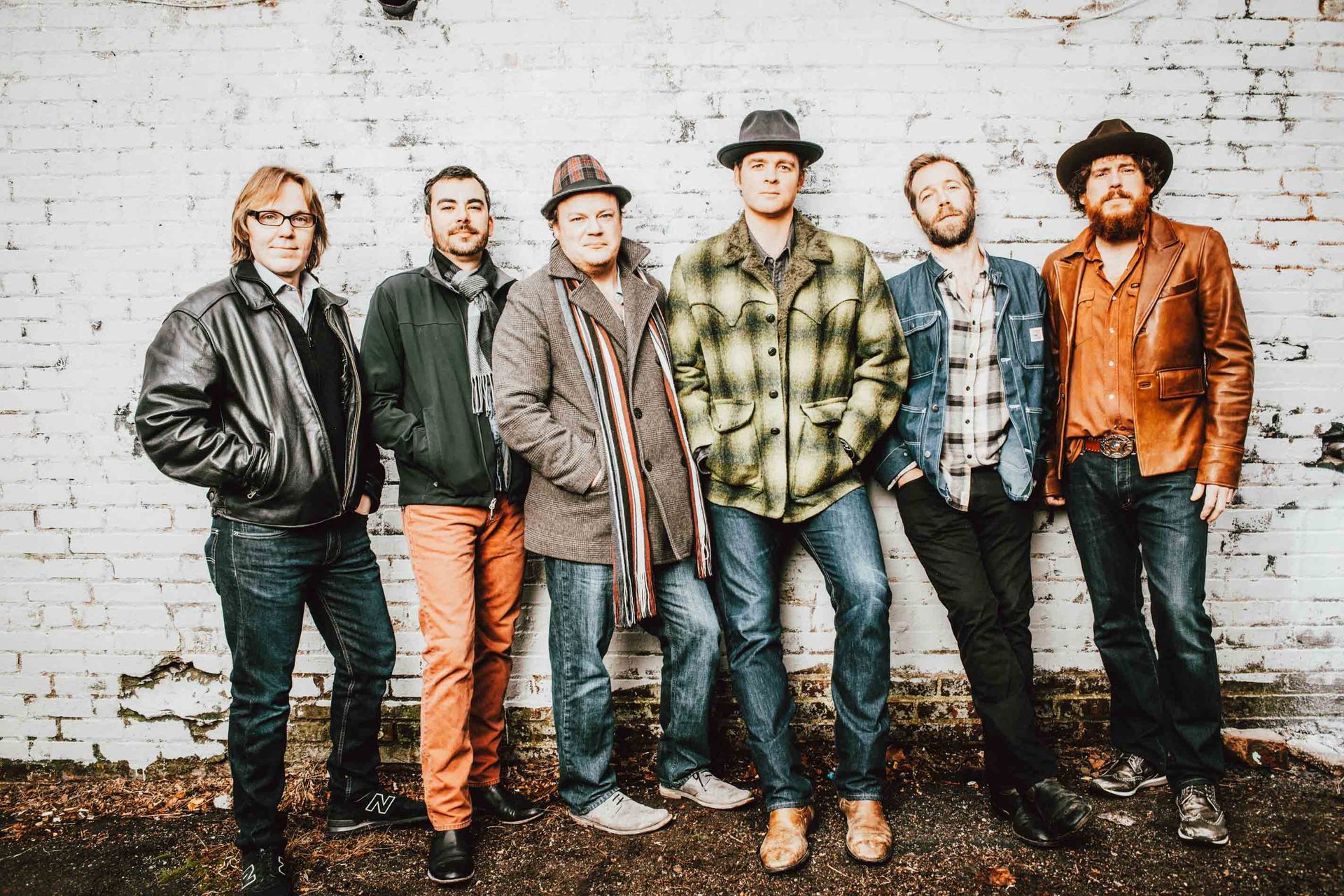 bluegrass musical group steep canyon rangers  standing against a white brick wall, wearing casual vintage-style clothes.