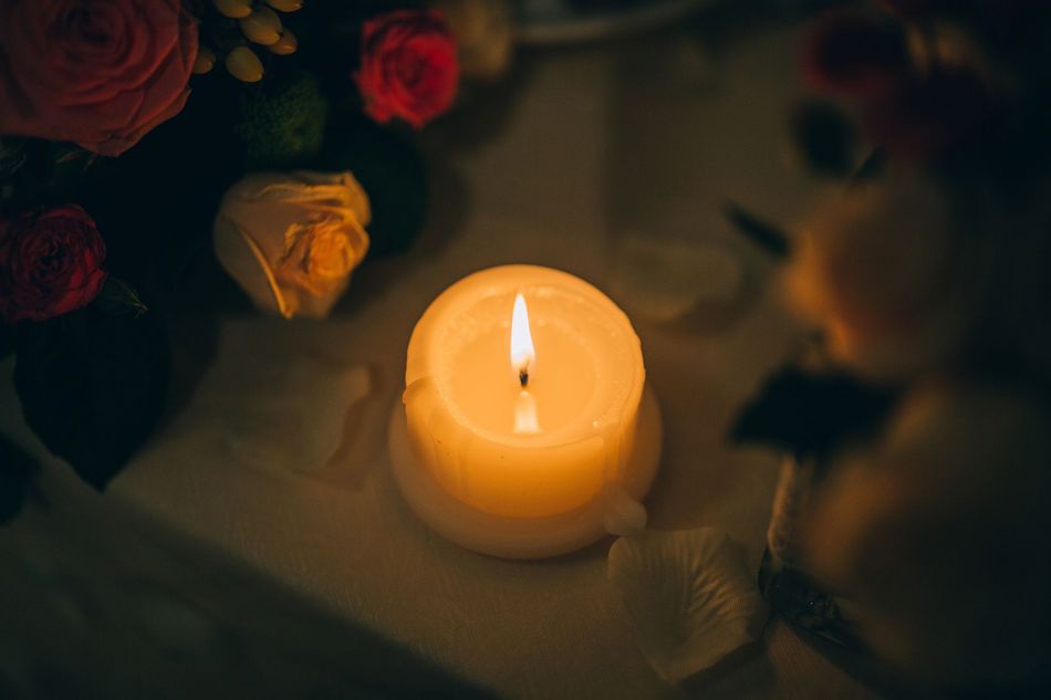 Lit white candle on a table, surrounded by soft-focus flowers in warm, dim light.