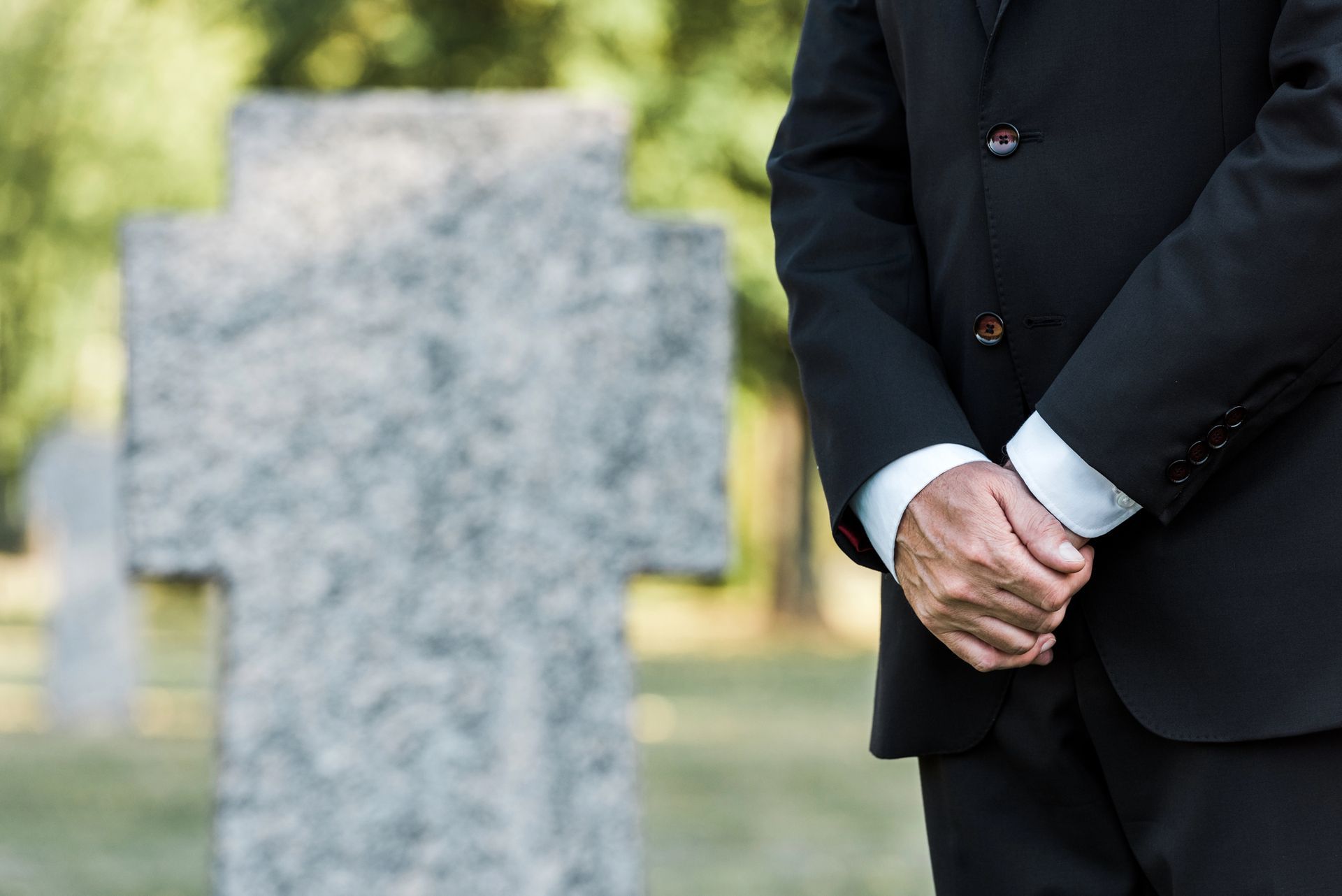 Mournful man in a black suit stands beside a gravestone, hands clasped in a cemetery.