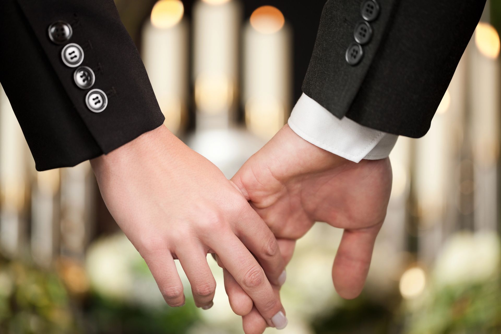 Two people in black suits holding hands in front of a blurred wedding ceremony backdrop