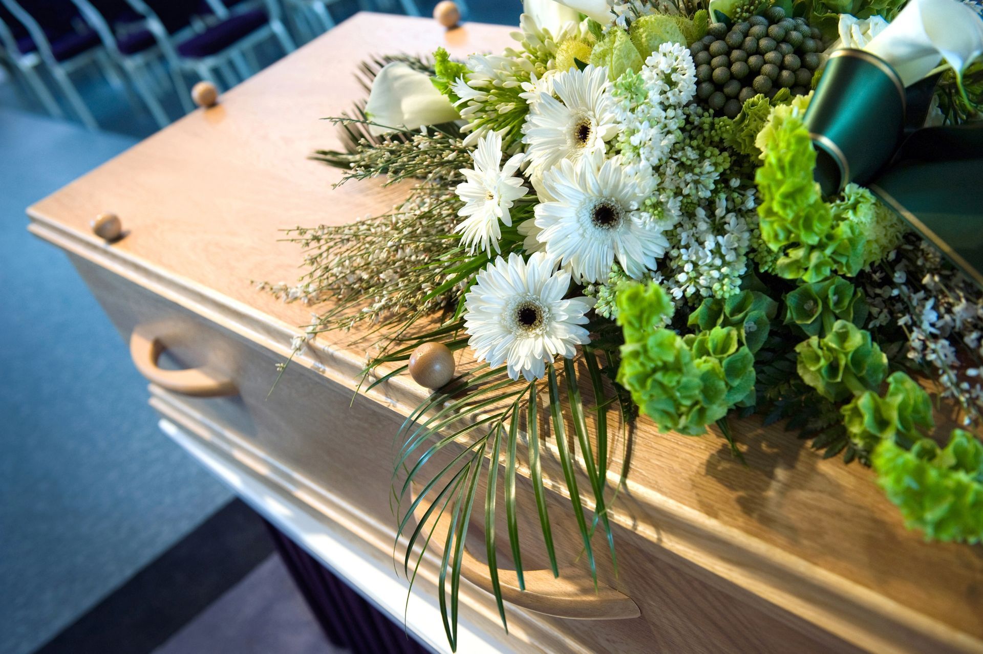 Wooden casket adorned with white flowers and green foliage at a funeral setting
