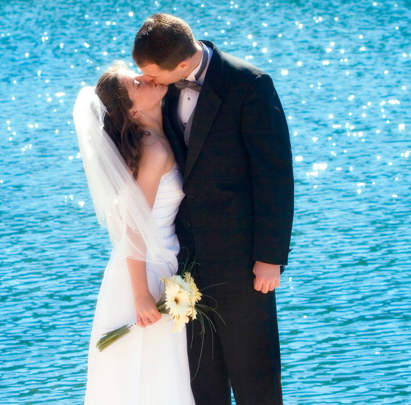 Bride and groom kissing near shimmering blue water; bride in white gown, holding flowers; groom in black suit.