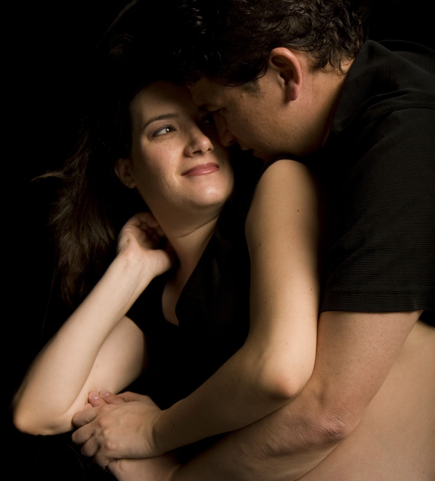 Couple embracing, softly lit; man's face near woman's, holding her arms. Woman smiles. Dark background.