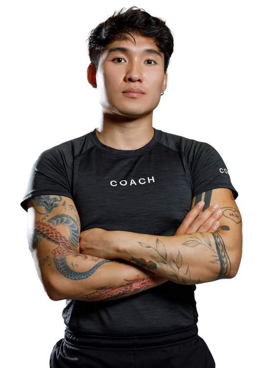 A coach in a dark athletic shirt with tattooed arms stands with arms crossed against a white background.