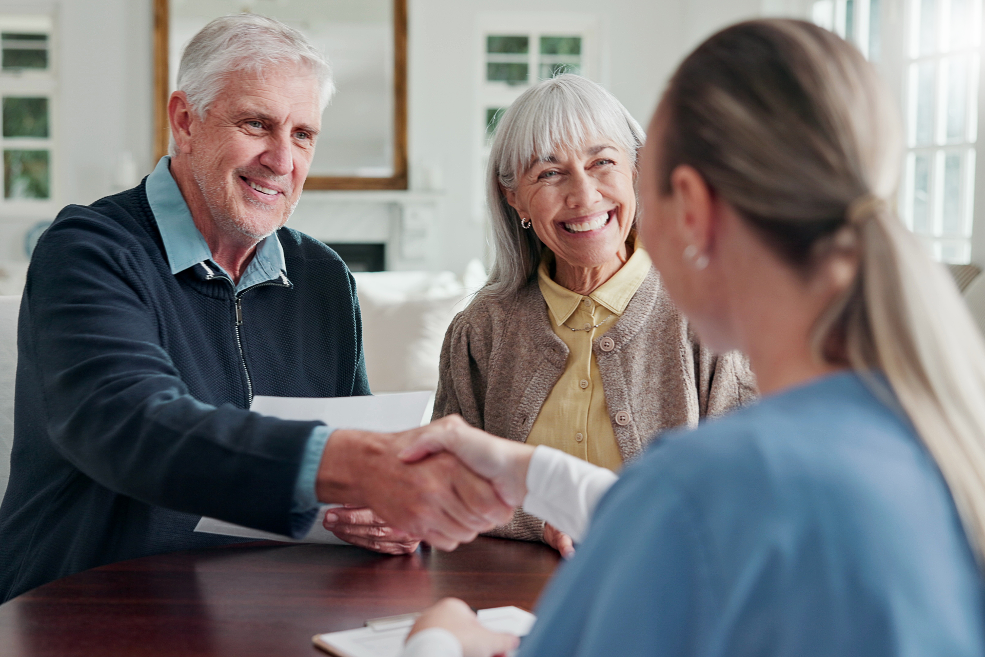Two people shaking hands across a table while a smiling woman sits nearby in a bright room