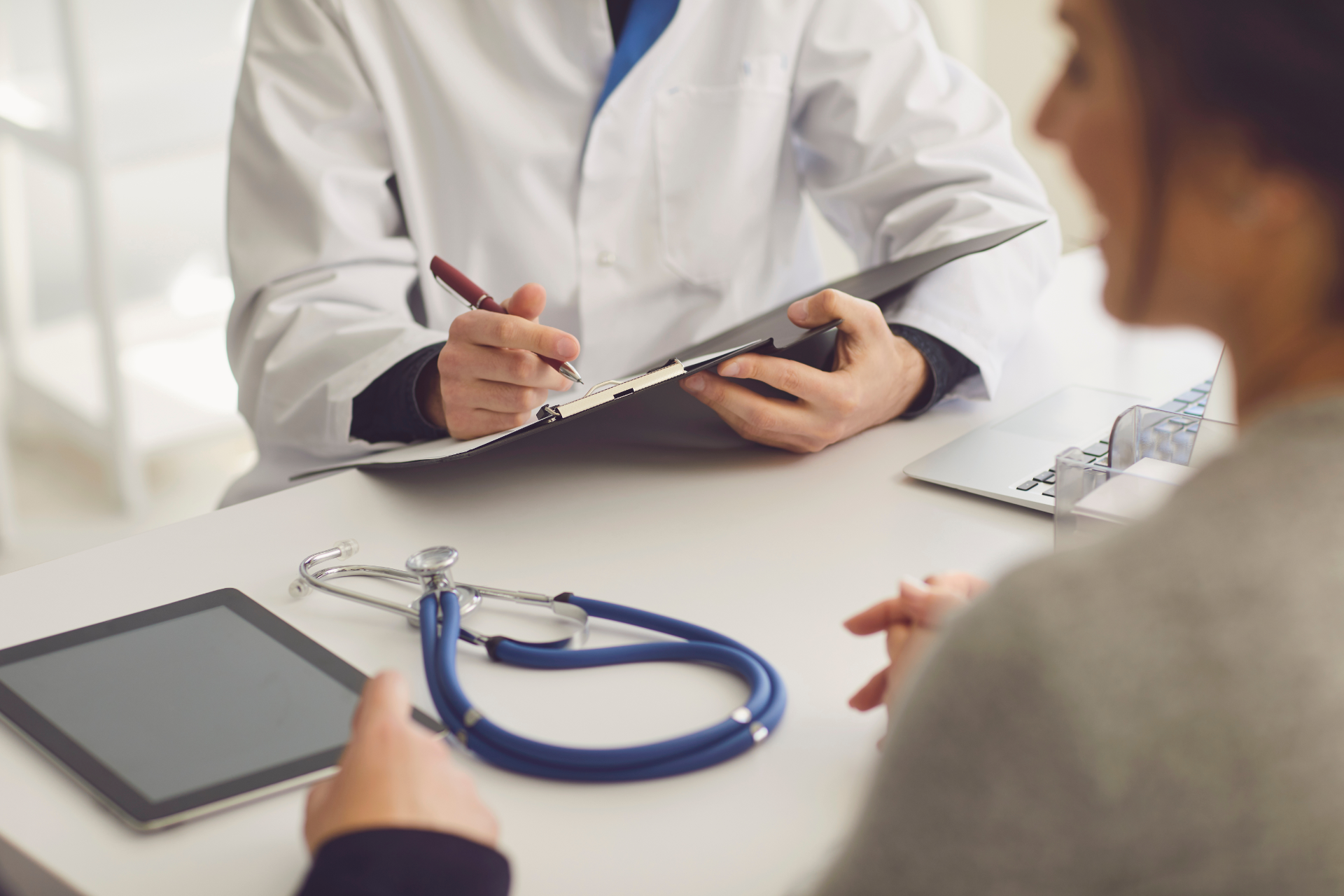 Doctor taking notes during a patient consultation with a stethoscope and tablet on the desk