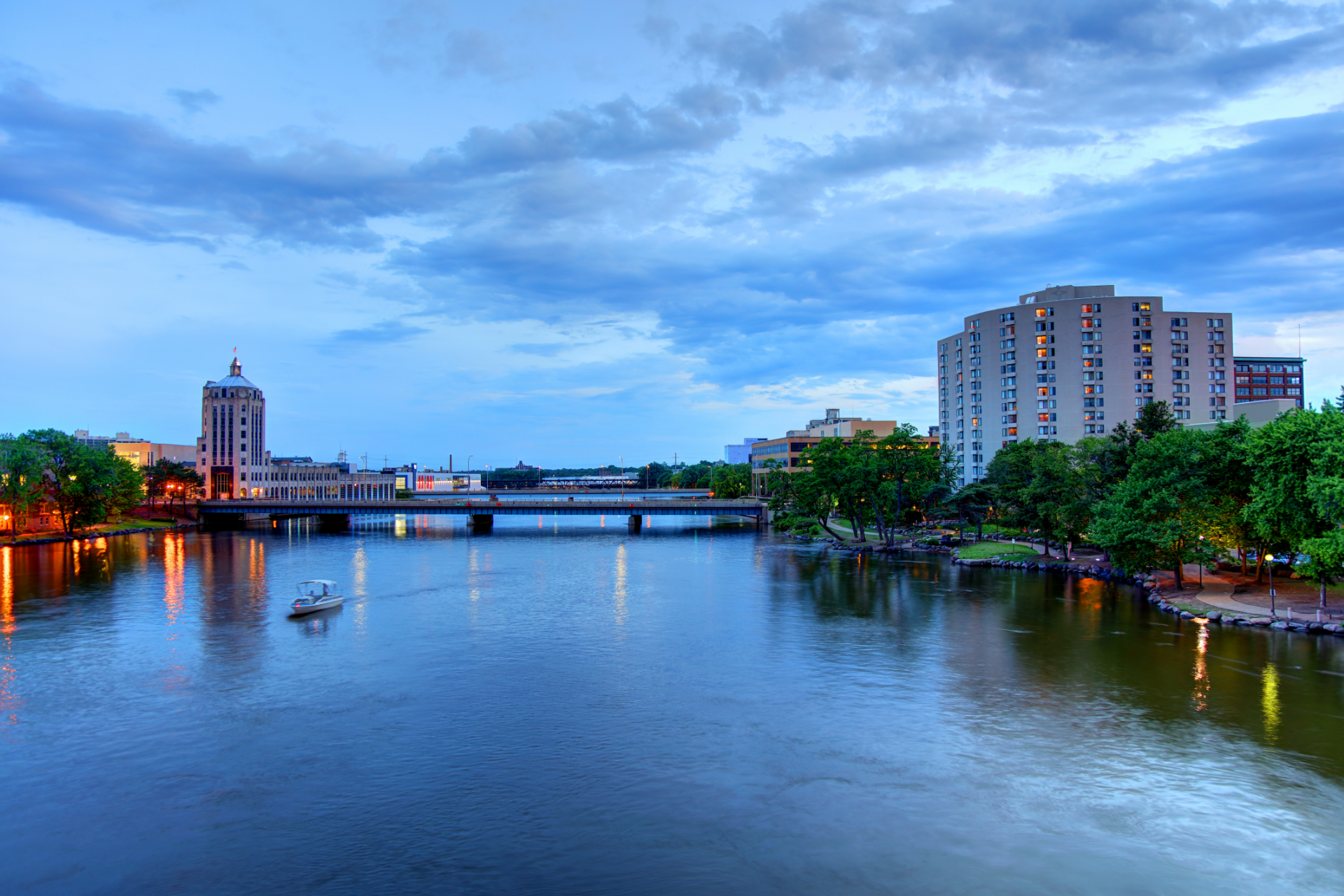River cityscape at dusk with a bridge, calm water, and lit buildings under a cloudy blue sky