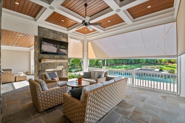 Outdoor living room with seating, fireplace, and pool view, partially shaded with a beige blind.