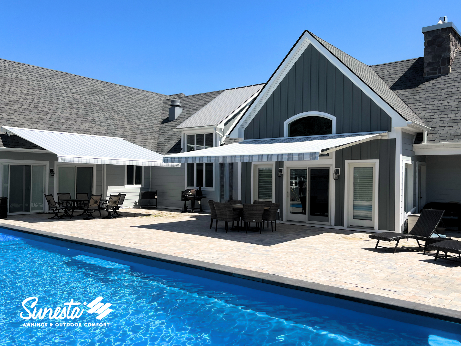 Swimming pool and patio with awnings at a house; blue water, blue and white awnings, gray siding.