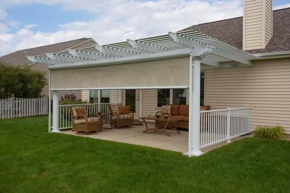 White pergola with beige shade over a concrete patio with outdoor furniture, next to a house with a beige siding.