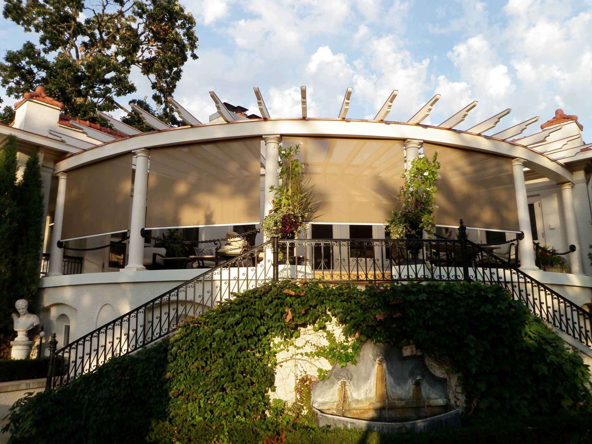Elegant white building with curved facade, awnings, and trellises. Lush greenery and stairs in the foreground.