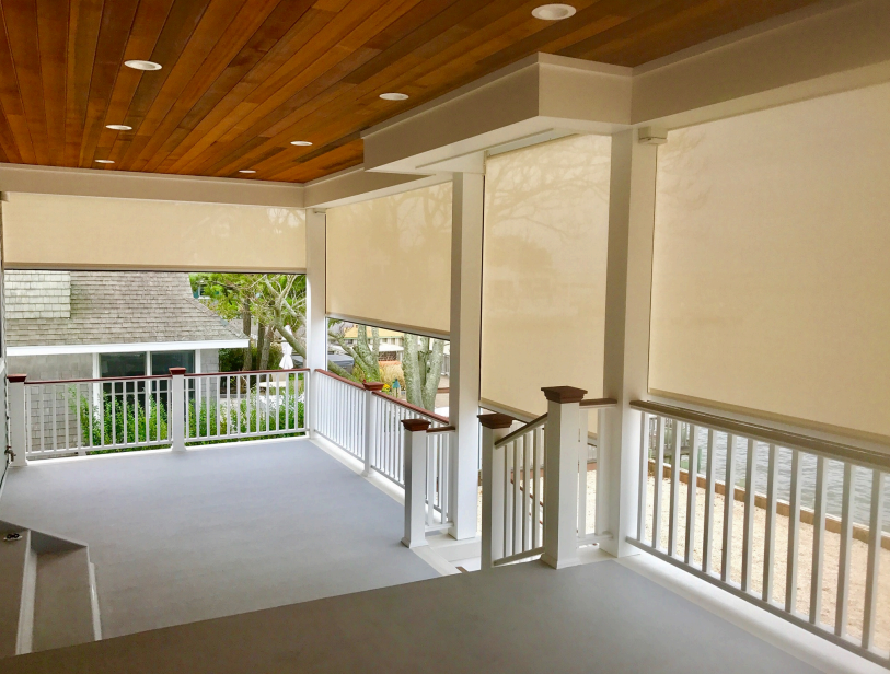A porch with white railings, retractable cream-colored shades, and a wooden ceiling.