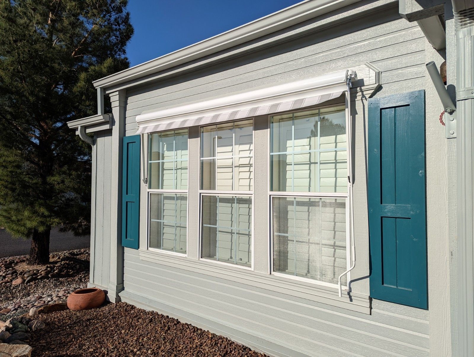 Gray house with teal shutters, a white awning, and three windows.