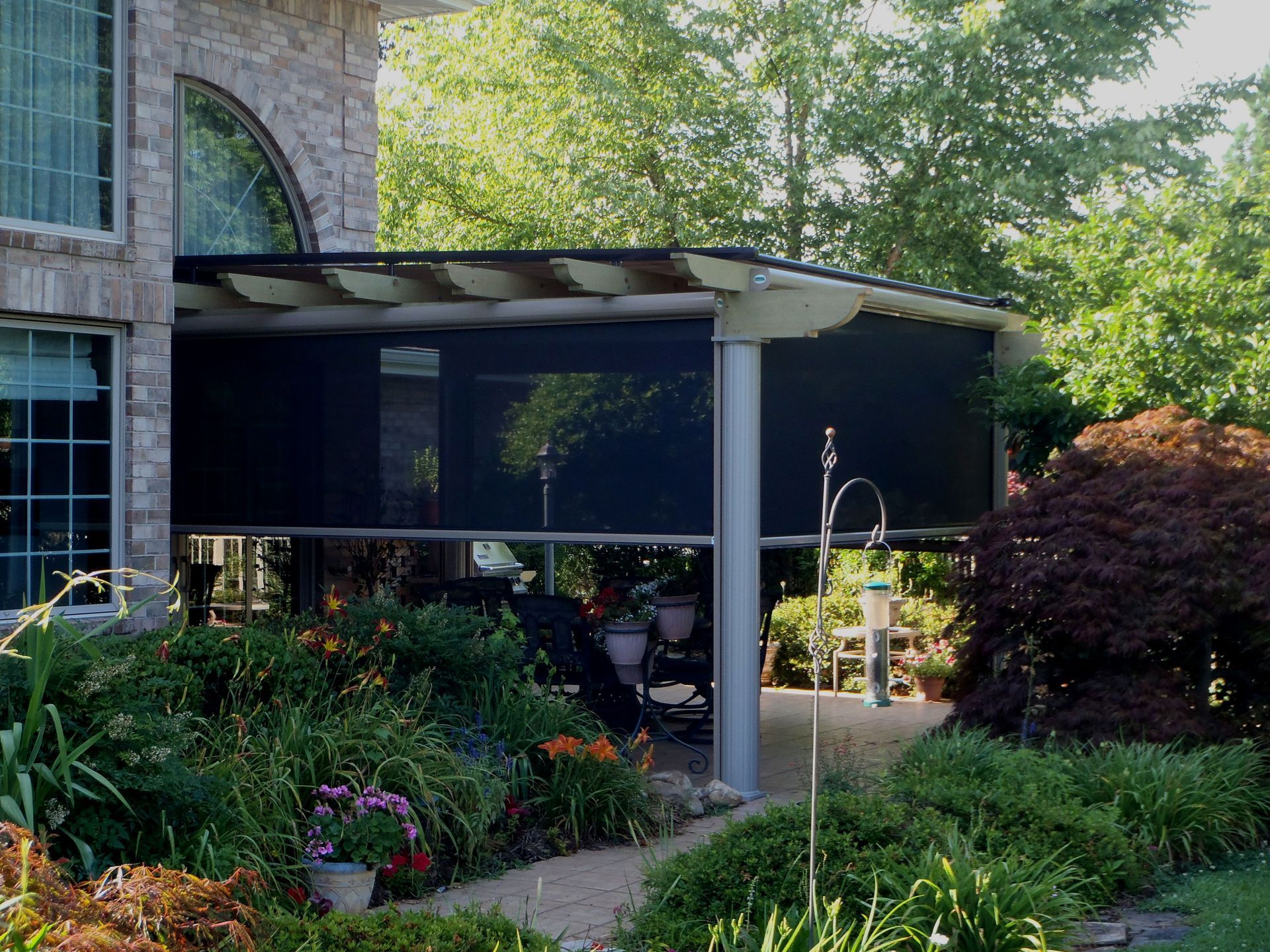 Covered patio with black sun shades next to a brick building, surrounded by lush garden.