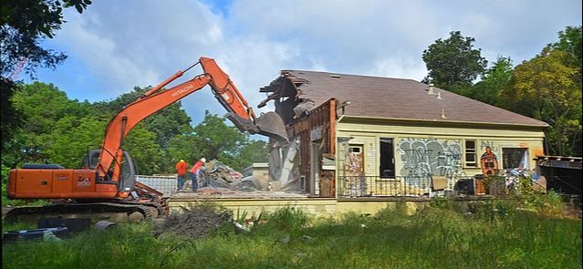 An excavator demolishes a building with graffiti-covered walls; workers in hard hats stand by.