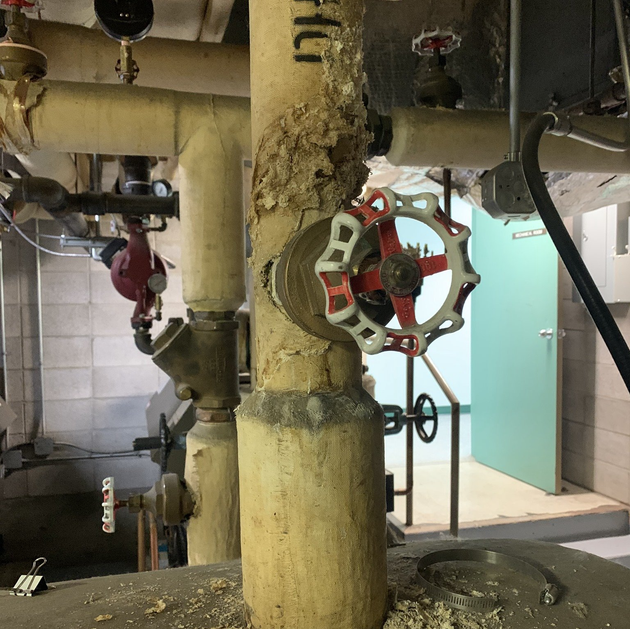 Pipes with insulation and valves in an industrial setting. A large red-and-white valve handle is visible.