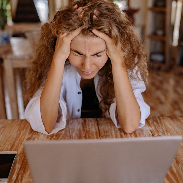Woman with curly hair, hands on head, looking at a laptop with a frustrated expression at a wooden table.