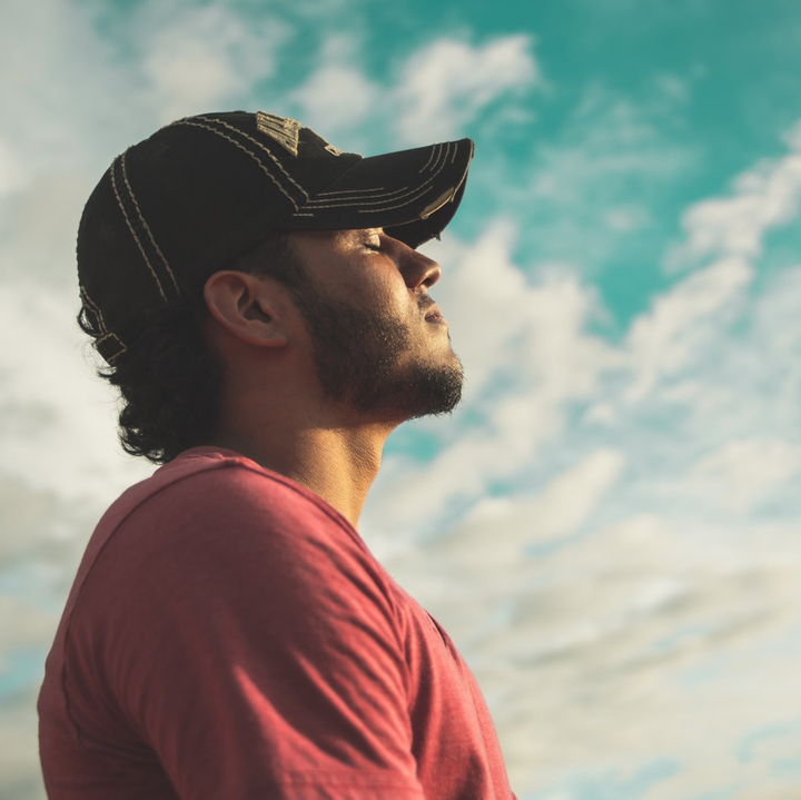 Man with a cap looking up at a blue sky with clouds, eyes closed.