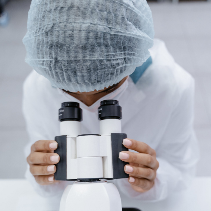 Person in lab coat and cap looking into a microscope in a laboratory setting.