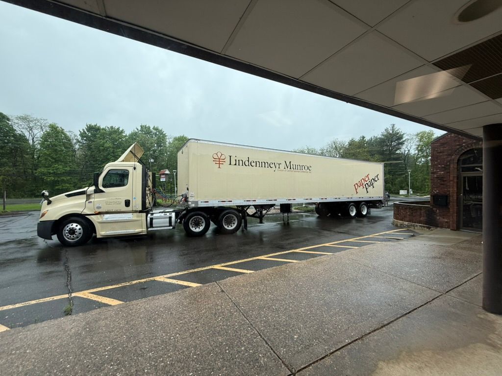 A beige semi-truck parked by a brick building on a wet, gray day; the trailer says