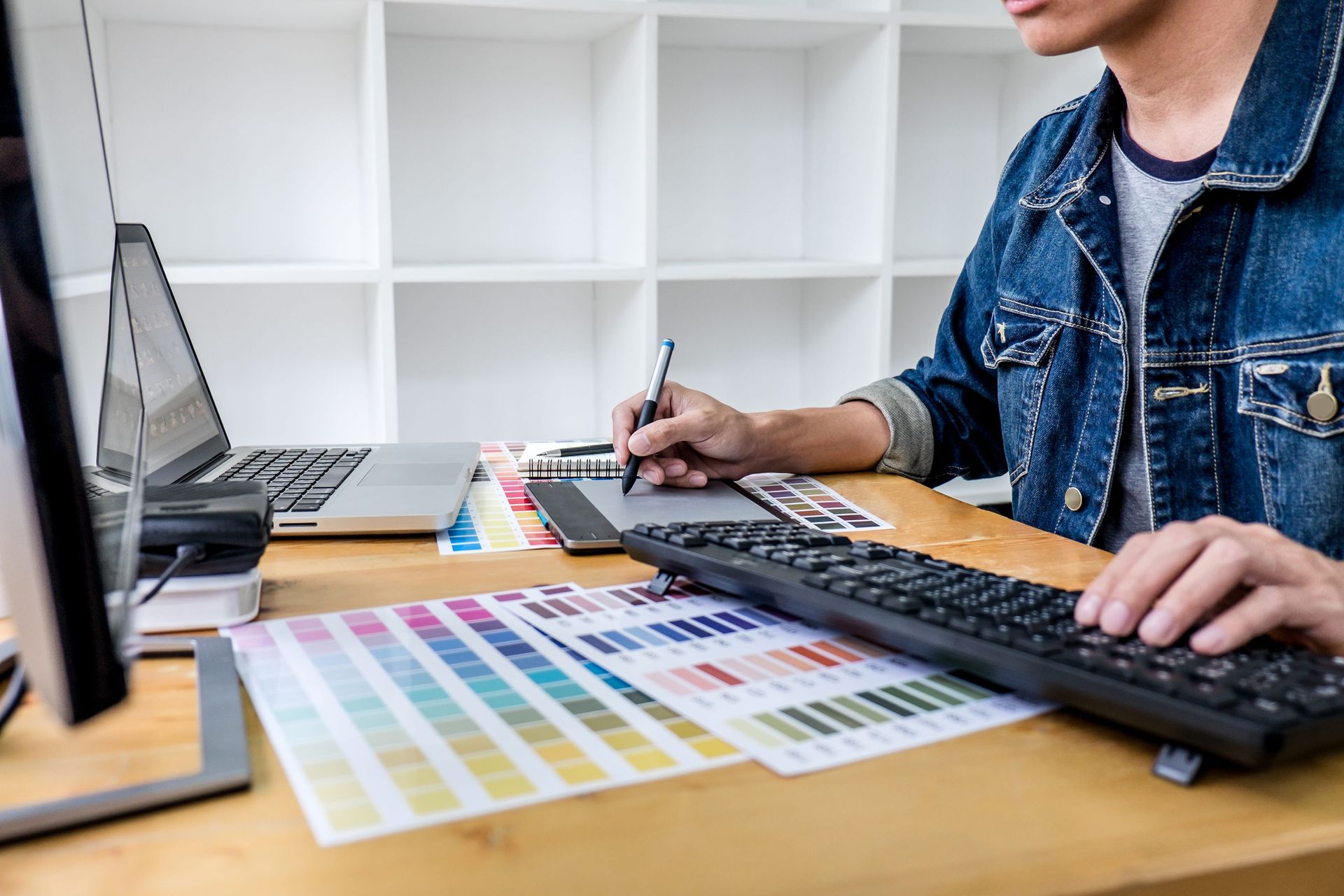 Person working at a desk with a keyboard, laptop, color swatches, and design samples.