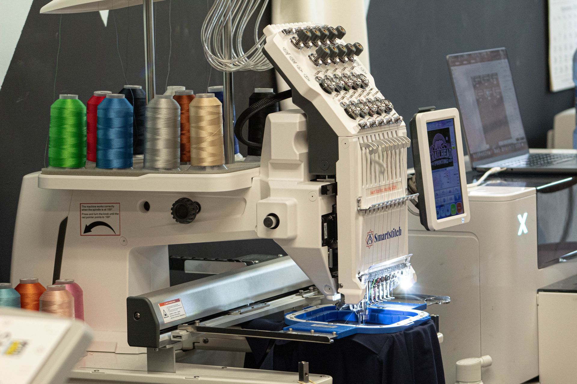 Embroidery machine stitching dark fabric, with colorful thread spools and a blue-lit work area.