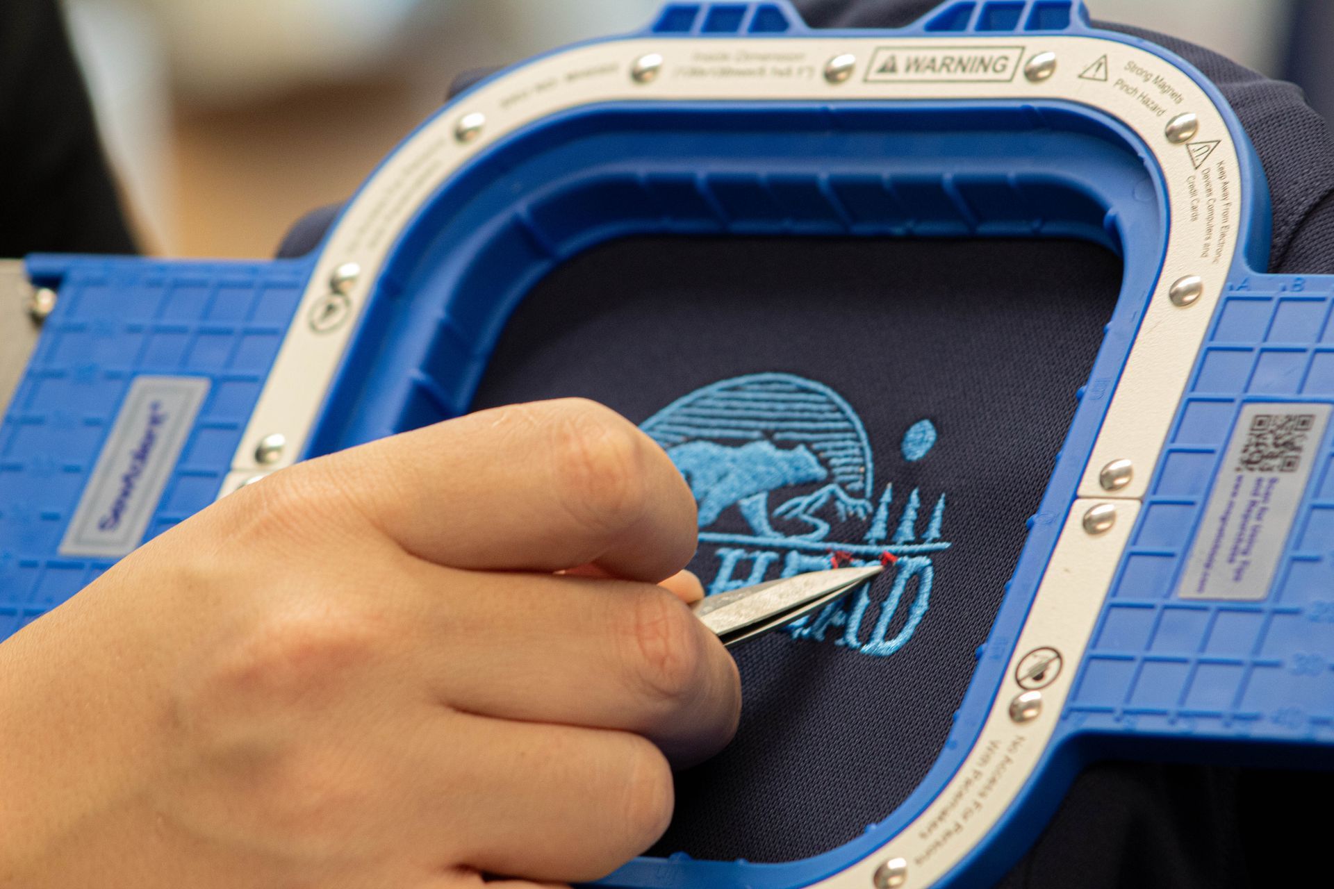 Embroidery machine stitching dark fabric, with colorful thread spools and a blue-lit work area.