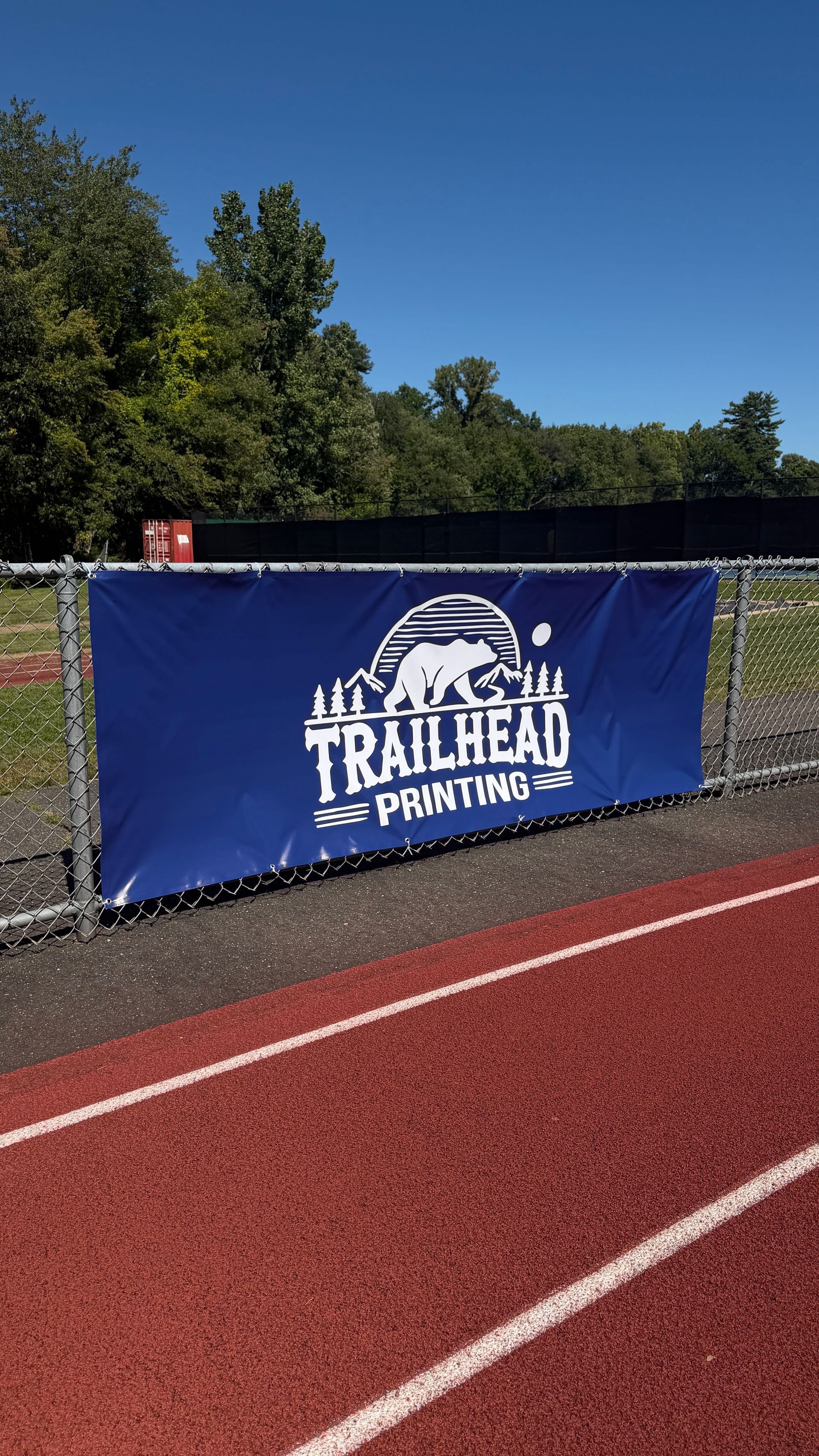 Blue Trailhead Running banner beside a red track under a clear sky, with trees in the background.