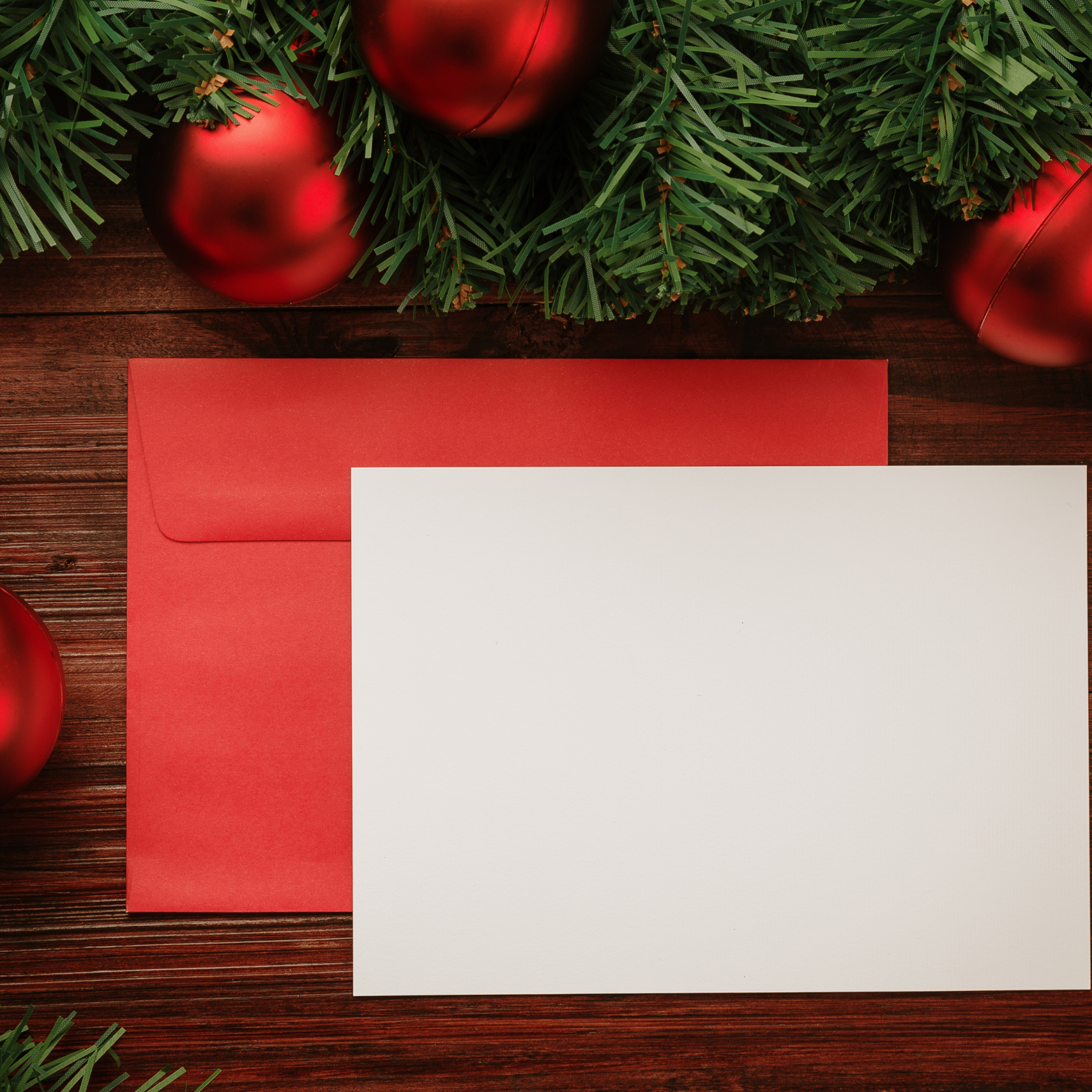Blank card and red envelope on a wooden surface with Christmas ornaments and greenery.