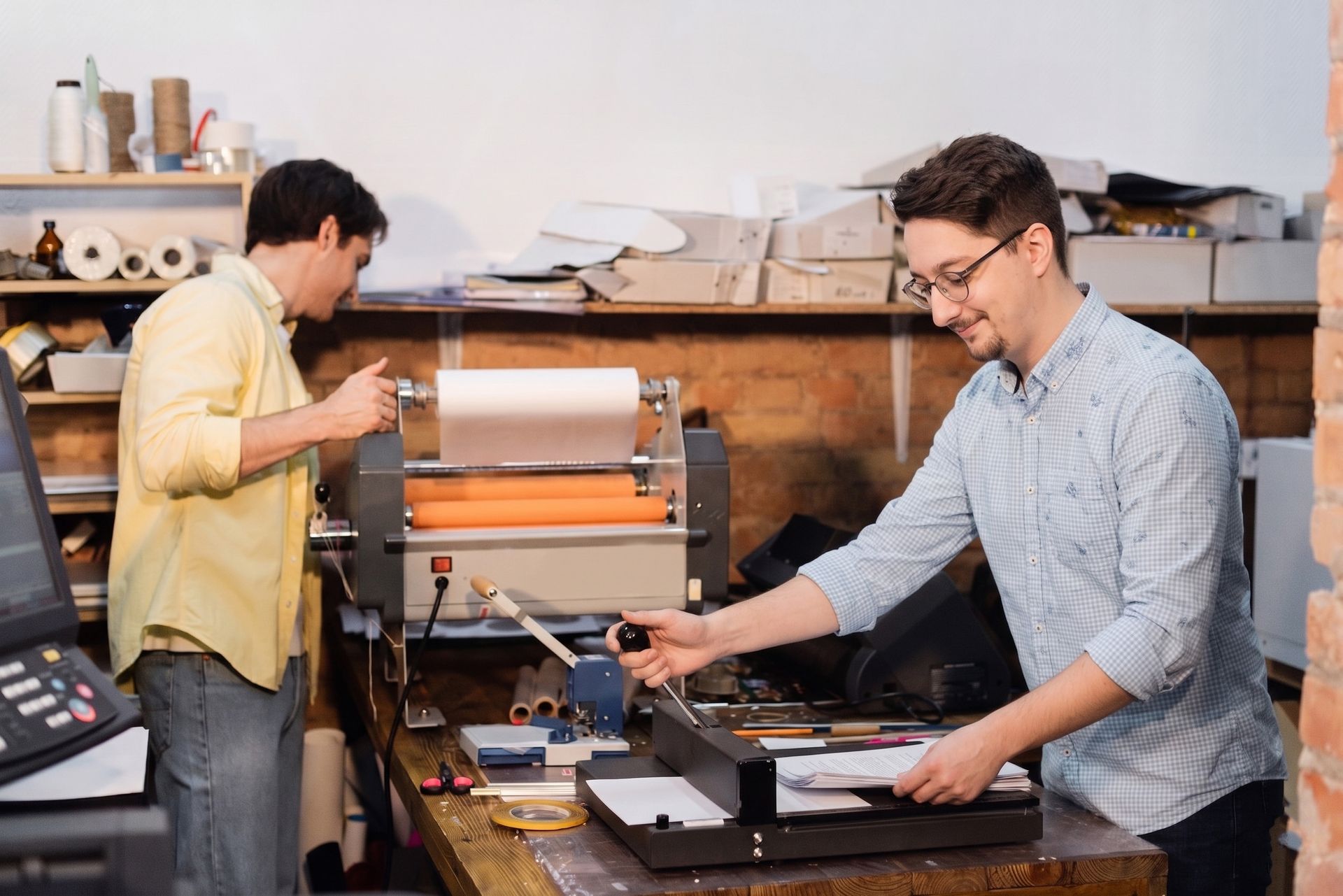 Two people working at a cluttered print shop counter, one adjusting a machine and the other loading paper