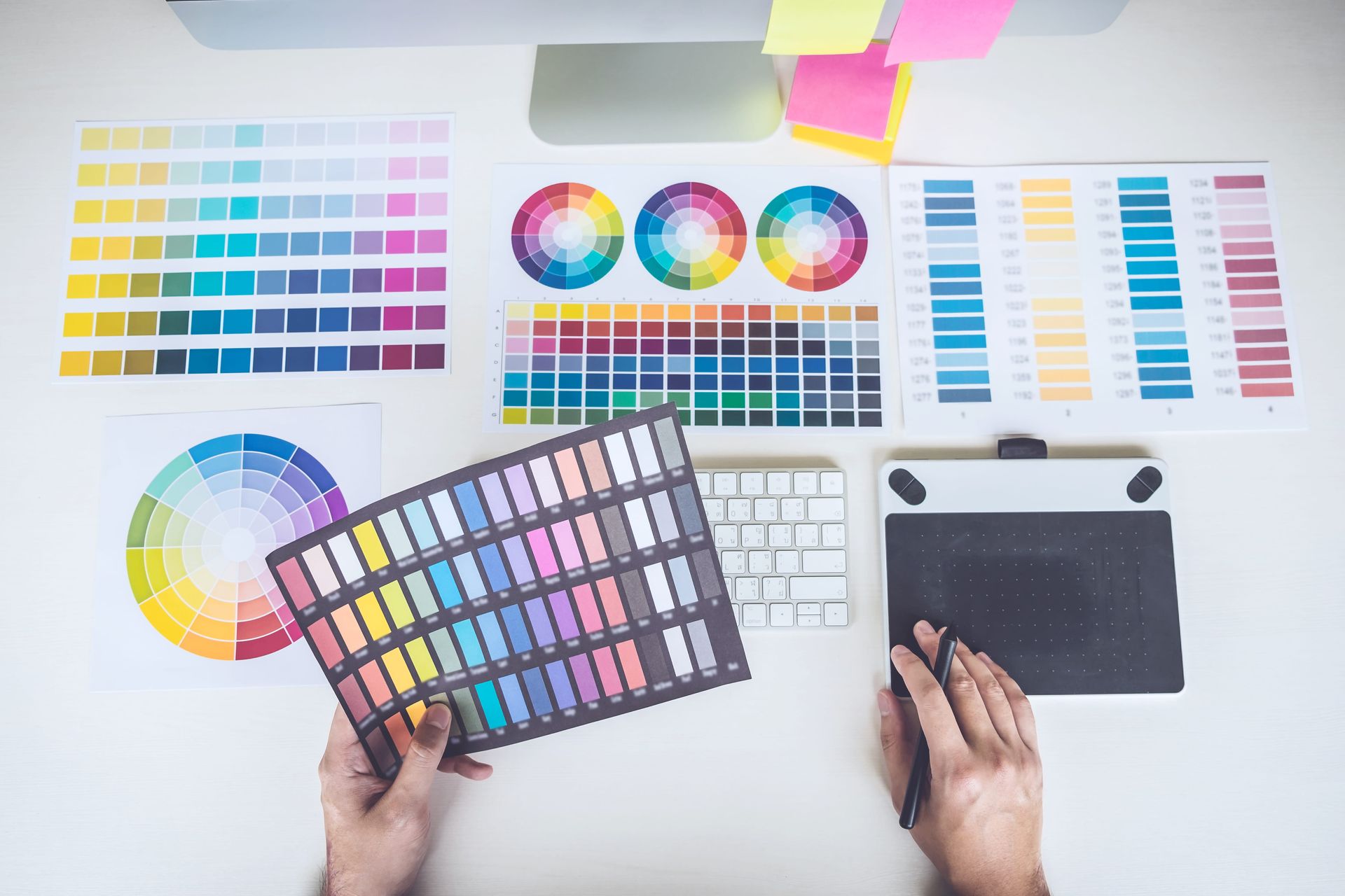 Hands holding color swatches and a tablet on a desk with color charts and palettes