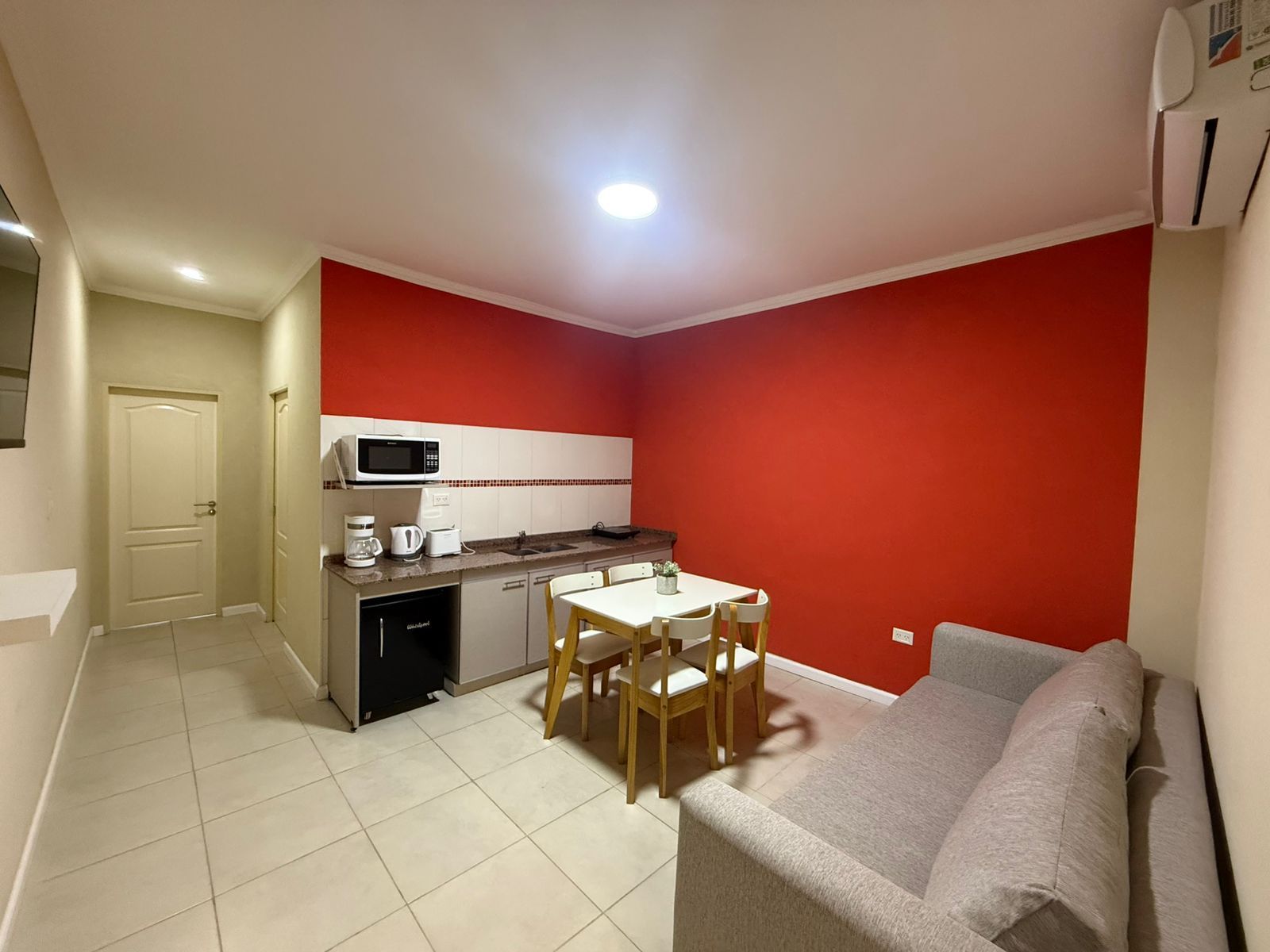 Interior de apartamento pequeño con cocina, mesa de comedor y sofá; paredes rojas y beige.