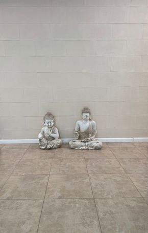Dos desgastadas estatuas de Buda se encuentran sobre un suelo de baldosas contra una pared de color beige.