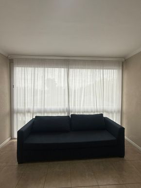 Sofá azul oscuro frente a cortinas blancas transparentes que cubren una ventana, paredes beige.