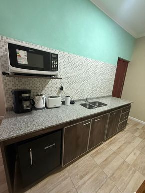 Cocina con microondas, cafetera y mininevera frente a un protector contra salpicaduras moteado y una pared verde azulado.