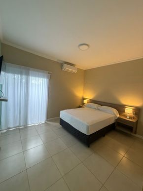 Dormitorio con cama, mesitas de noche y puerta corredera con cortinas transparentes. Paredes claras y suelo de baldosas.
