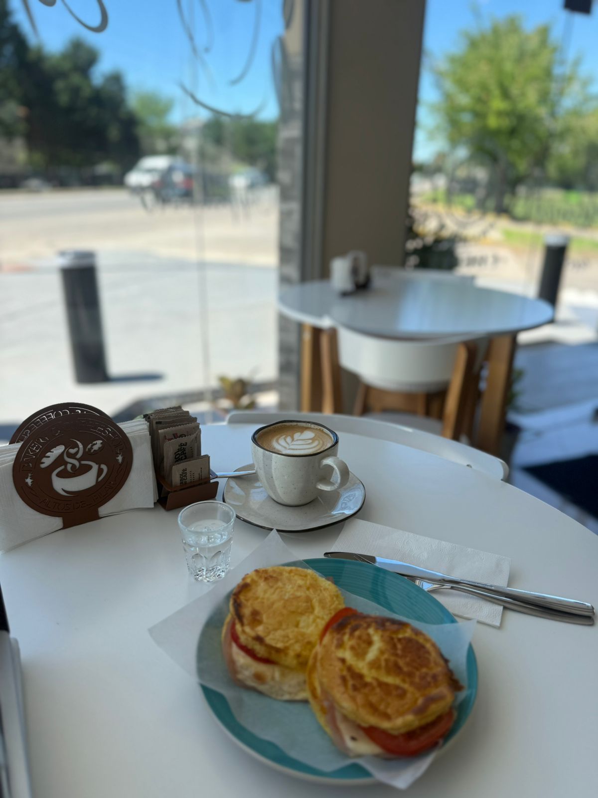 Desayuno en una cafetería: café, sándwich de huevo y agua en una mesa blanca cerca de una ventana.