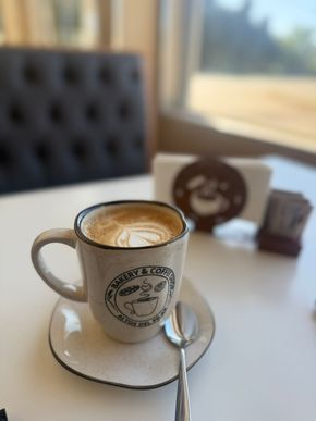 Latte en una taza moteada sobre un plato con cuchara, sobre una mesa blanca. Fondo borroso de un restaurante.