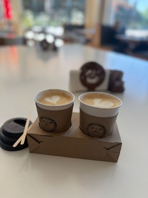 Dos cafés con leche en vasos de papel con arte en una bandeja, junto a tapas y agitadores, sobre una mesa blanca.
